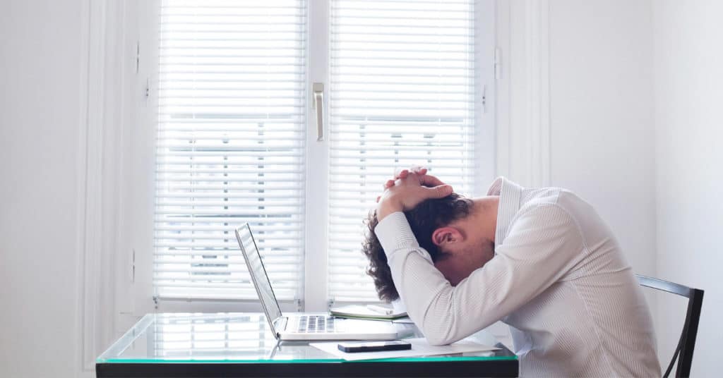 man in white at desk with head down on table due to stress